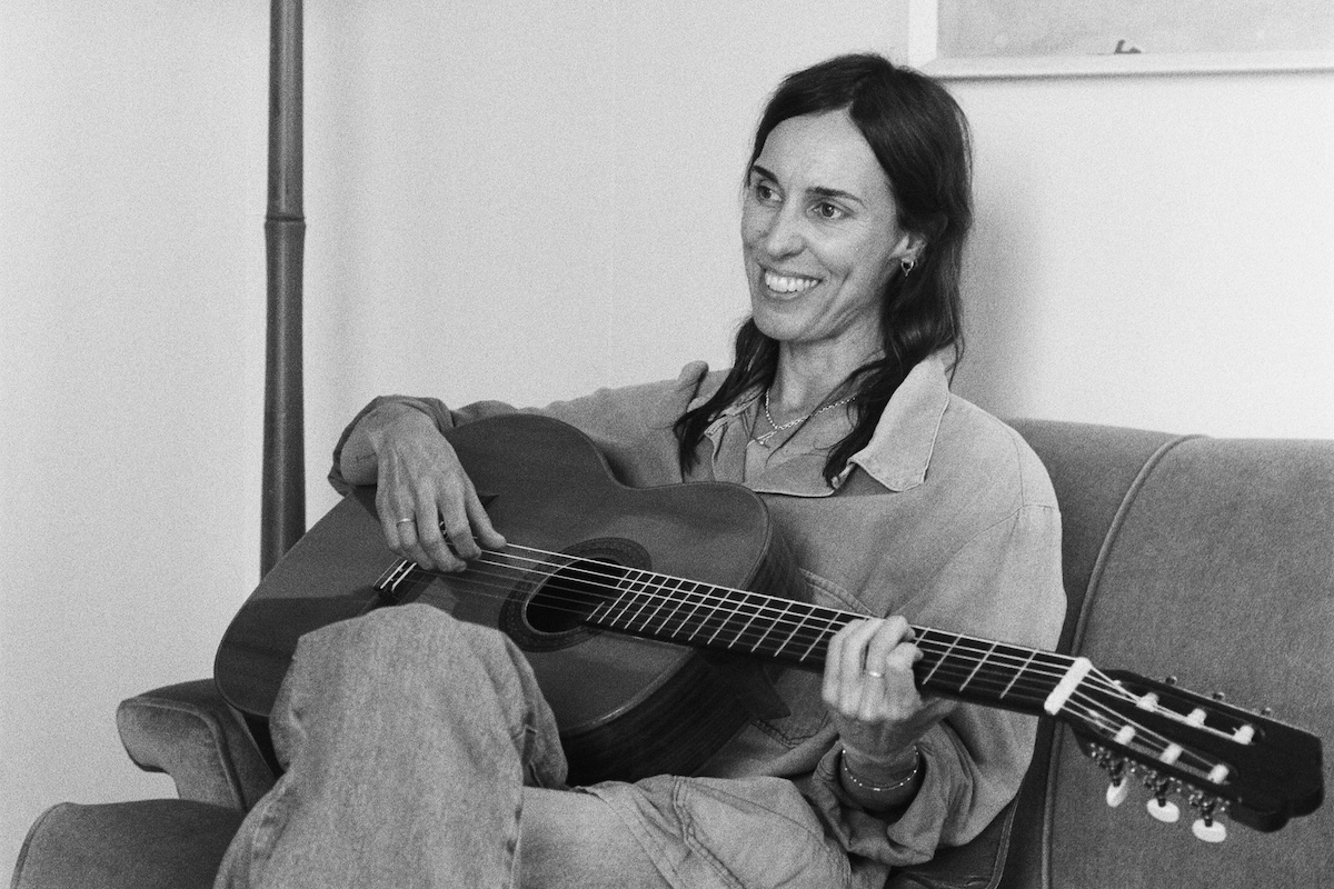 Black and white photo of Australian musician Holly Throsby smiling while playing acoustic guitar indoors on a couch.